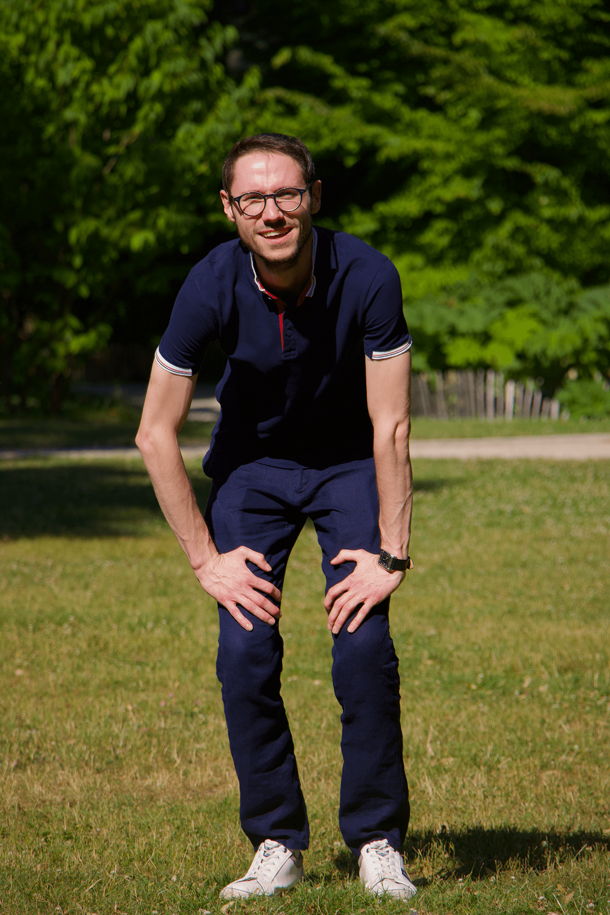 Man in a navy polo and pants bending forward on a grassy park lawn, smiling at the camera, wearing glasses.