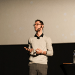 Male presenter on a stage, wearing glasses and a headset mic, gesturing with his hands during a talk.
