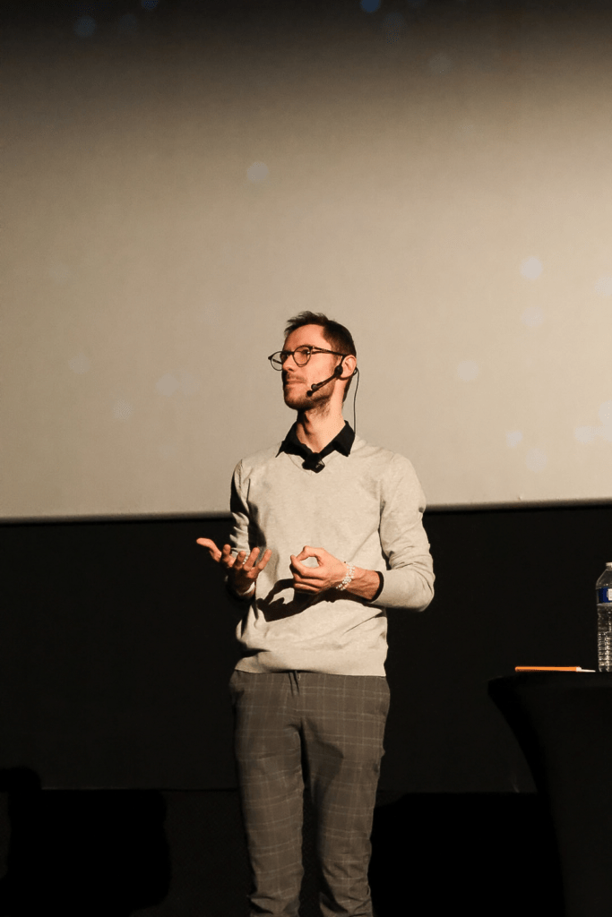 Male presenter on a stage, wearing glasses and a headset mic, gesturing with his hands during a talk.