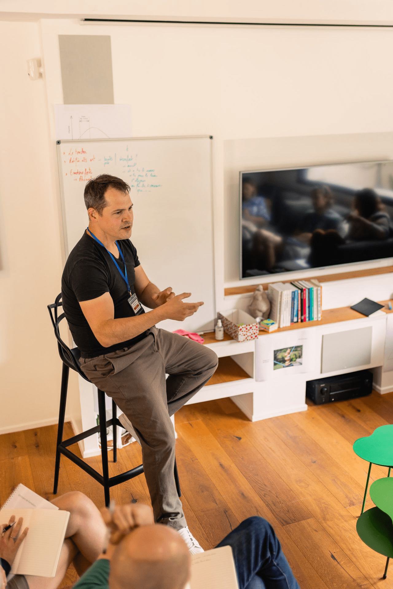Male presenter in a black T‑shirt sits on a tall stool, gesturing as he teaches a group taking notes in a bright room with a whiteboard and a wall-mounted TV behind him.